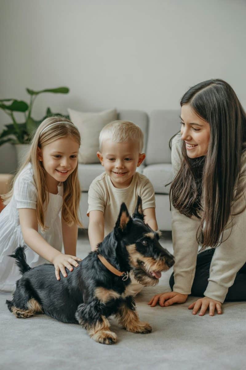 Three people - a young girl with long blonde hair, a young boy with short light-colored hair, and a young woman with long dark hair - sitting on a light gray floor, interacting with a black and tan terrier mix dog.