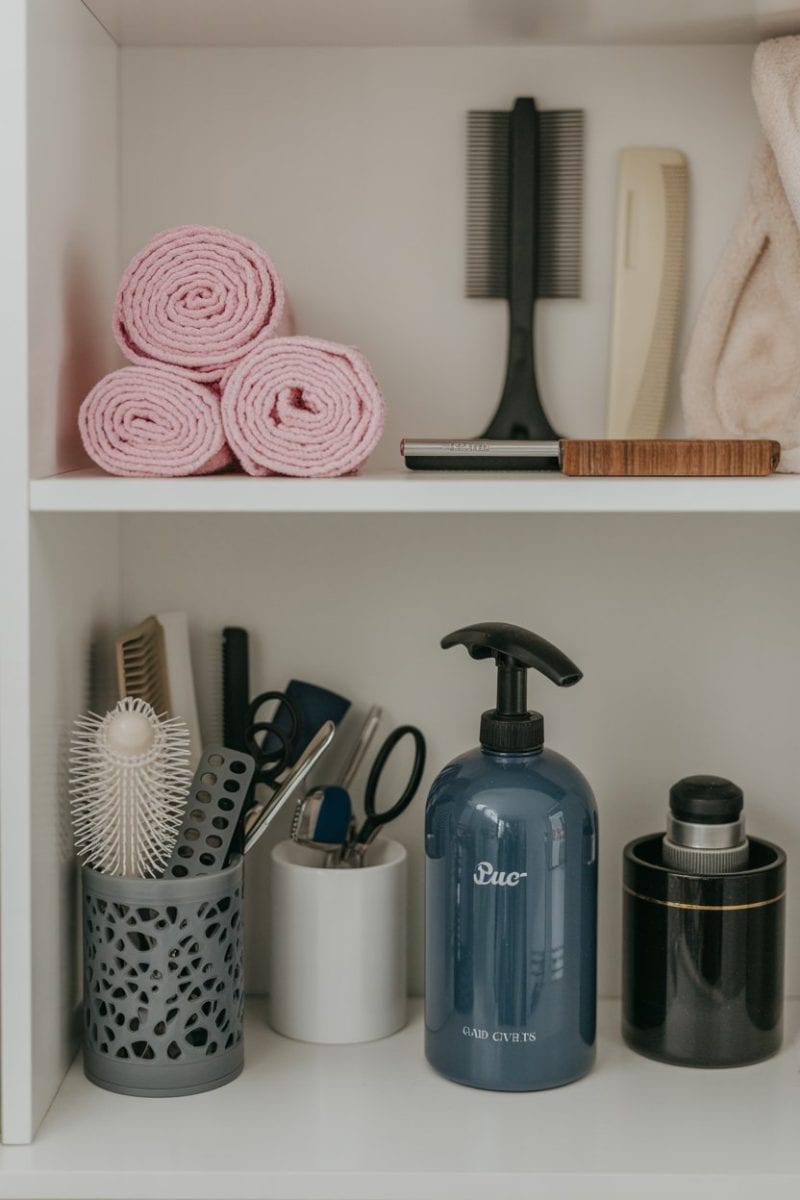The top shelf contains three rolled pink towels stacked together, and a black wooden-handled hair brush with bristles. The bottom shelf displays various personal care items: a black mesh container holding hair brushes and scissors, a white cylindrical container with small grooming tools.