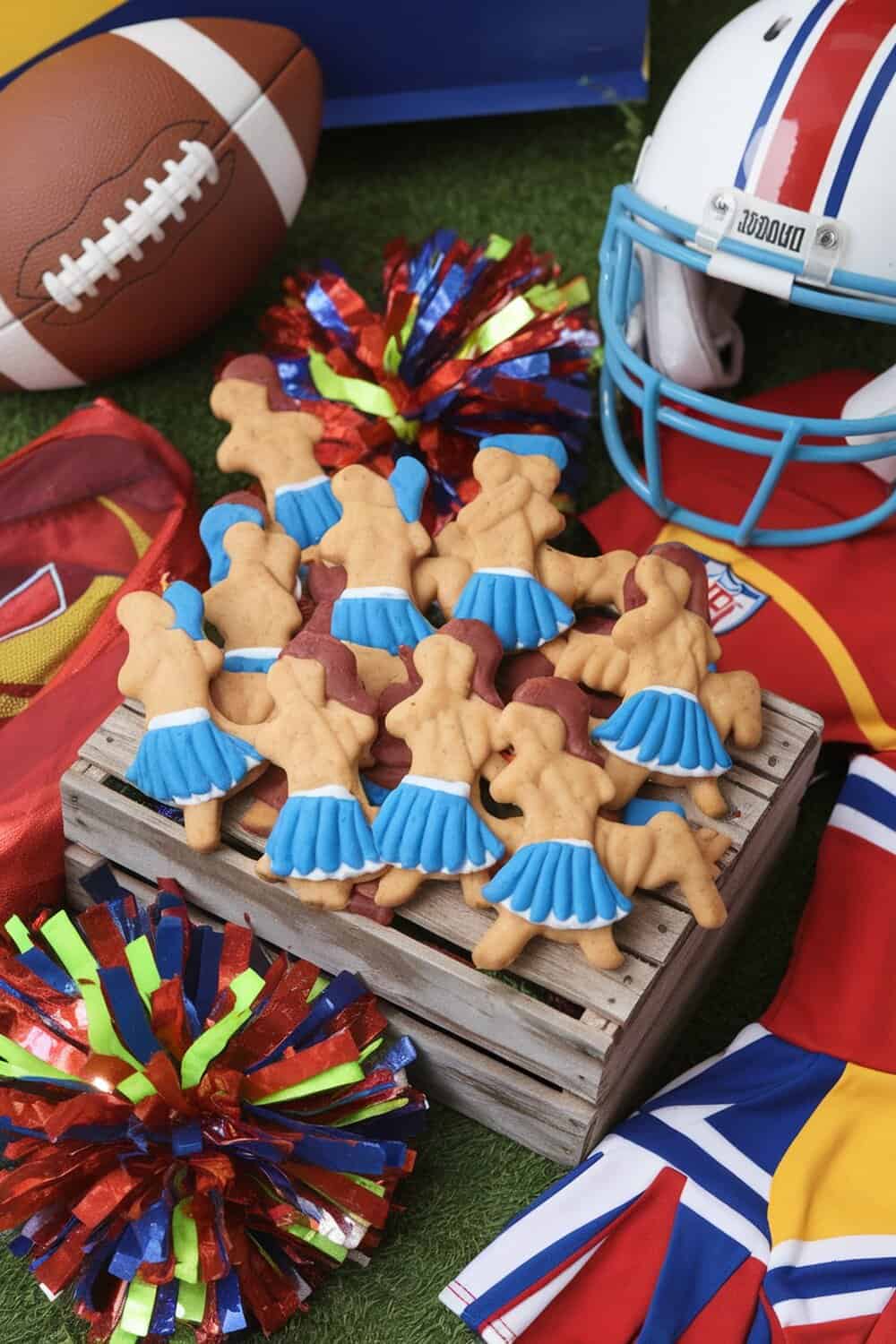 A pile of cheerleader-shaped dog treats with blue skirts, surrounded by football gear and pom-poms.